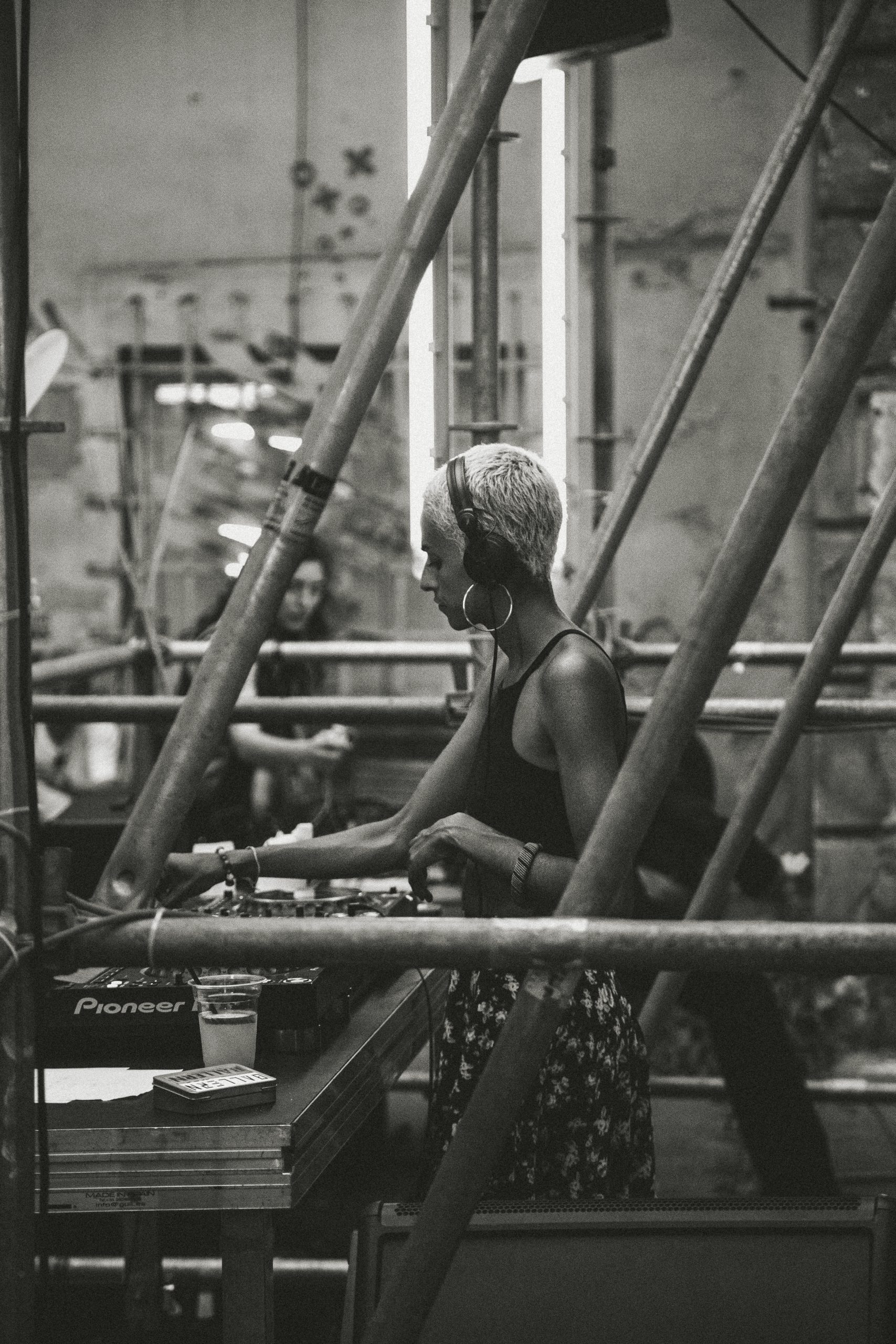 DJ artist with headphones and very short hair is dj-ing on an industrial deck with metal poles.